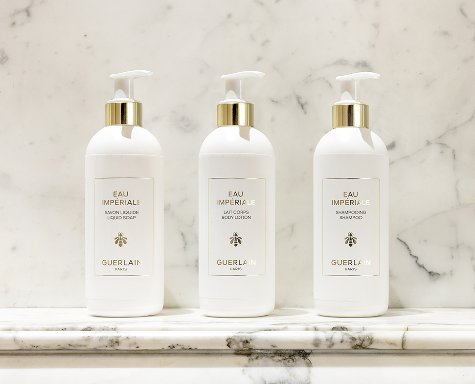 Três frascos brancos de Guerlain , rotulados como sabonete líquido, loção corporal e champô, expostos sobre uma superfície de mármore.