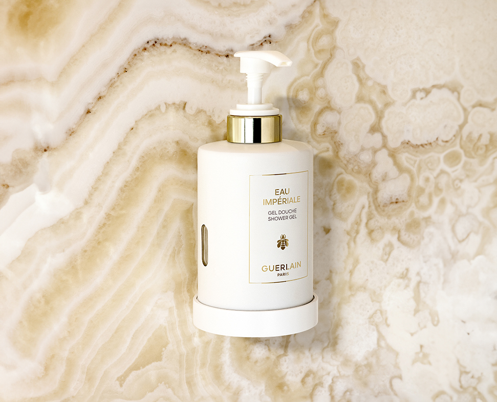 Um frasco branco com bomba do gel de banho Guerlain Imperiale é colocado contra um fundo de mármore texturizado. O frasco apresenta um detalhe dourado e um rótulo com o nome do produto.