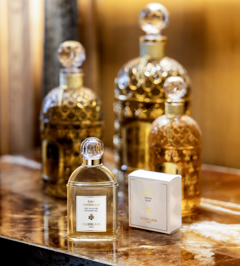 Uma coleção de elegantes frascos de perfume, incluindo um com a etiqueta «Eau Imperiale», expostos sobre uma superfície de mármore com iluminação suave ao fundo.
