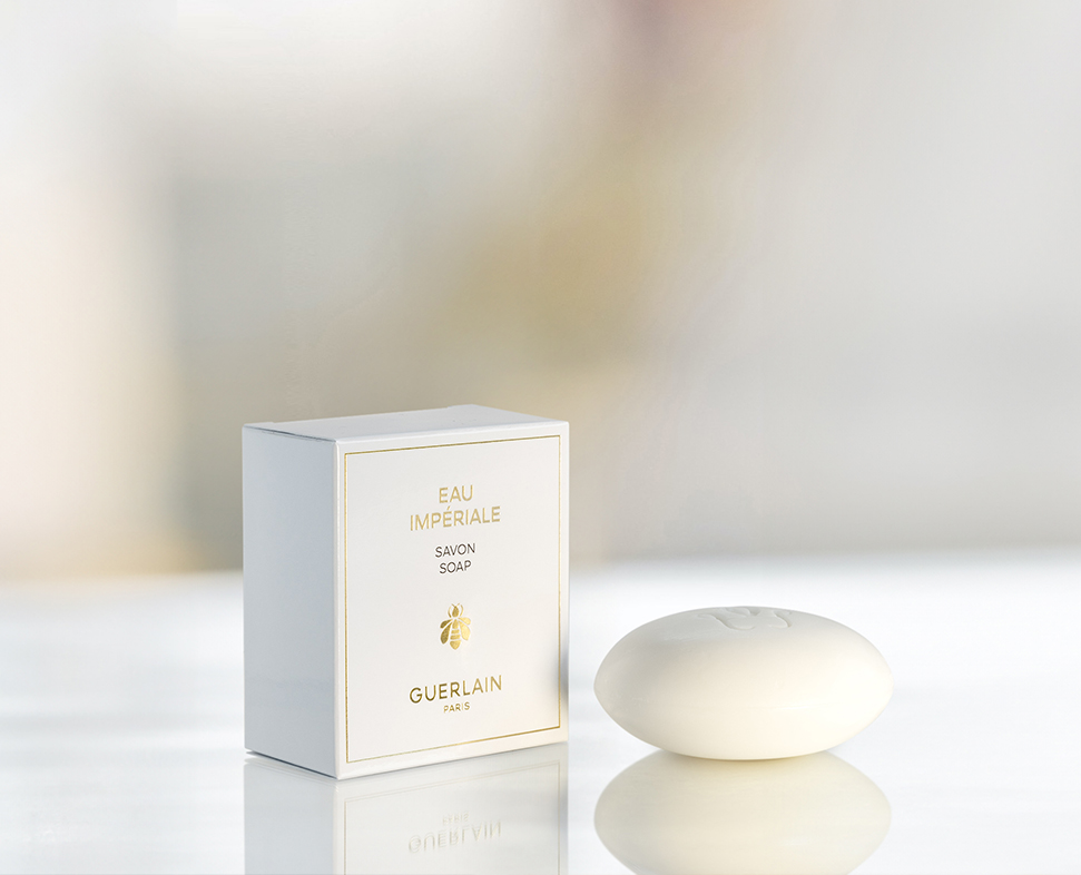 Uma barra de sabonete branca ao lado da sua elegante embalagem, com a etiqueta «Eau Impériale Salon Soap» da Guerlain, contra um fundo suave e desfocado.