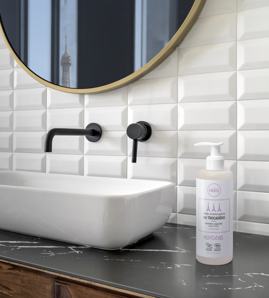 Un moderno lavabo da bagno con una vasca quadrata bianca e un rubinetto nero, caratterizzato da uno specchio rotondo decorativo e una bottiglia di sapone Huygens sul ripiano, con la vista della Torre Eiffel riflessa nello specchio.