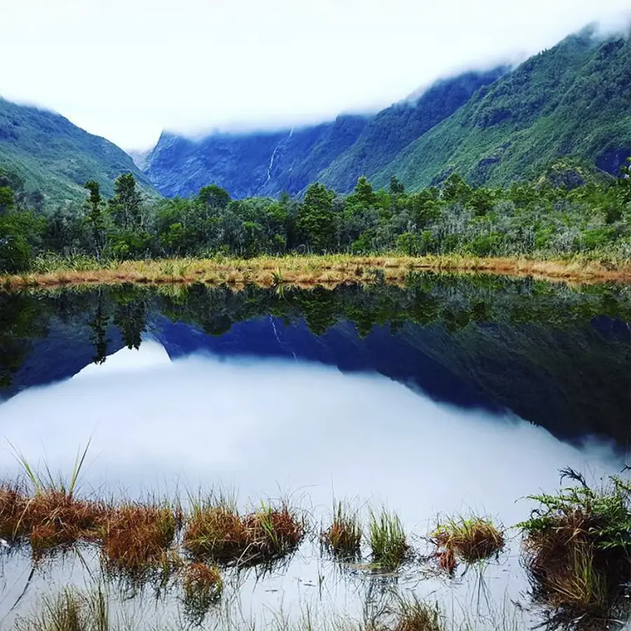 Un paesaggio sereno caratterizzato da un lago calmo che riflette le montagne circostanti e la vegetazione rigogliosa, con nuvole basse che contribuiscono a creare un'atmosfera nebbiosa.