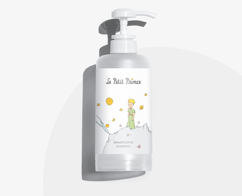Een shampoofles met pompdop, voorzien van een speelse illustratie van een jongetje, geïnspireerd op „De Kleine Prins“, omringd door sterren en planeten. Op het etiket staat „Le Petit Prince Shampooing.”