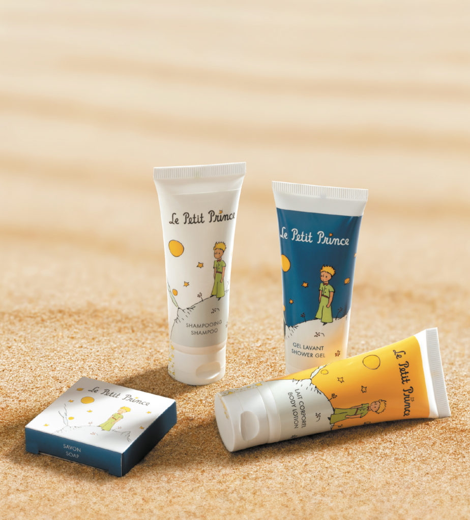Vier Körperpflegeprodukte mit Illustrationen ausLe Petit Prince, darunter ein Shampoo, ein Duschgel und ein Stück Seife, auf einer sandigen Oberfläche platziert.
