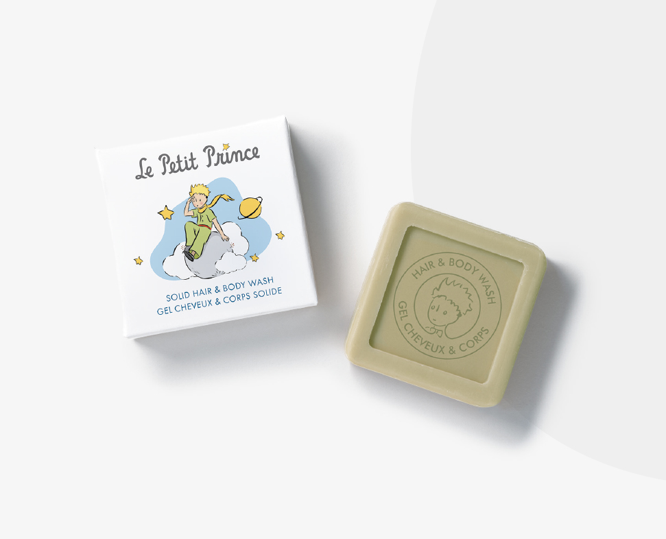 Ein festes Haar- und Körperwaschstück in einem grünen Behälter neben seiner Verpackung mit einer verspielten Illustration eines Kindes und Wolken, beschriftet mitLe Petit Prince“.