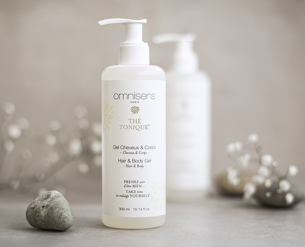 Ένα μπουκάλι Omnisens Hair and Body Gel, με την ένδειξη «Gel Cheveux &amp; Corps» και αντλία διανομής, τοποθετημένο σε ουδέτερο φόντο με απαλό φωτισμό και διακοσμητικές πέτρες.
