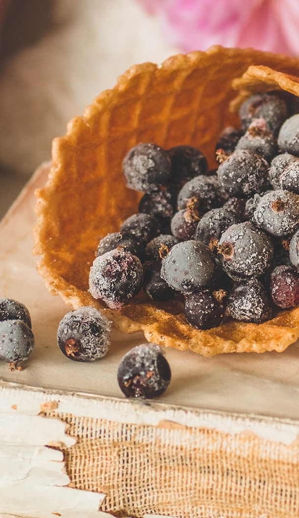 Eine mit gefrorenen Blaubeeren gefüllte Waffeltüte liegt auf einer strukturierten Oberfläche. Die Blaubeeren sind mit Raureif überzogen, was ihnen ein kühles Aussehen verleiht.