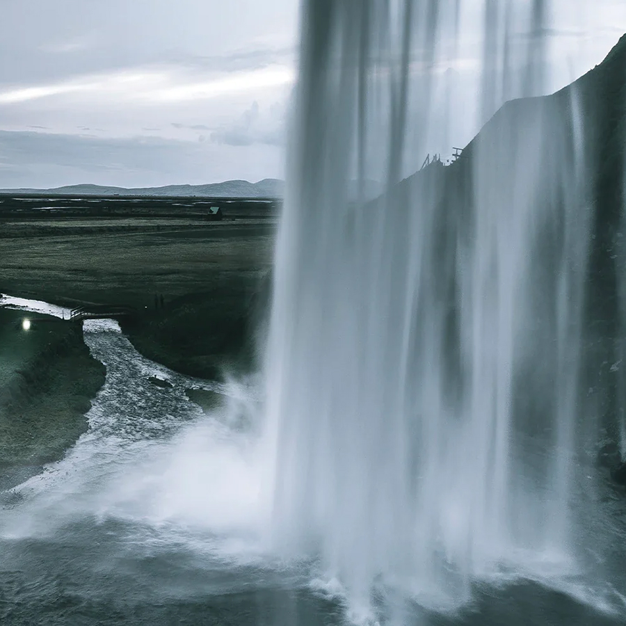 Un primo piano di una cascata che si riversa su una sporgenza rocciosa, con la nebbia che sale e un fiume che scorre più in basso. Il paesaggio presenta dolci colline sullo sfondo, sotto un cielo nuvoloso.