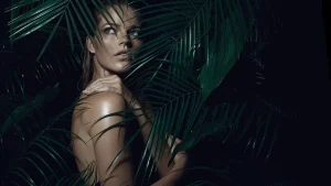 Una donna parzialmente nascosta è in piedi tra rigogliose foglie di palma verdi, con una luce soffusa che illumina il suo viso e il suo corpo. Guarda di lato, trasmettendo un senso di fascino e bellezza naturale.