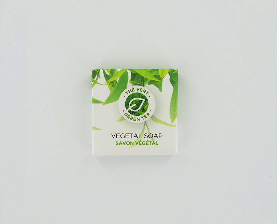Uma pequena caixa quadrada de sabonete vegetal com um desenho de chá verde e a inscrição «Vegetal Soap» em inglês e francês. O fundo é branco com detalhes em folhas verdes.