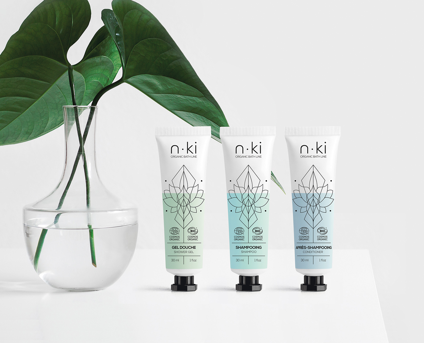 Drei Tuben mit n-ki Bio-Haarpflegeprodukten, darunter Shampoo und Gel, sind neben einer durchsichtigen Vase mit einer grünen Pflanze vor einem minimalistischen Hintergrund angeordnet.
