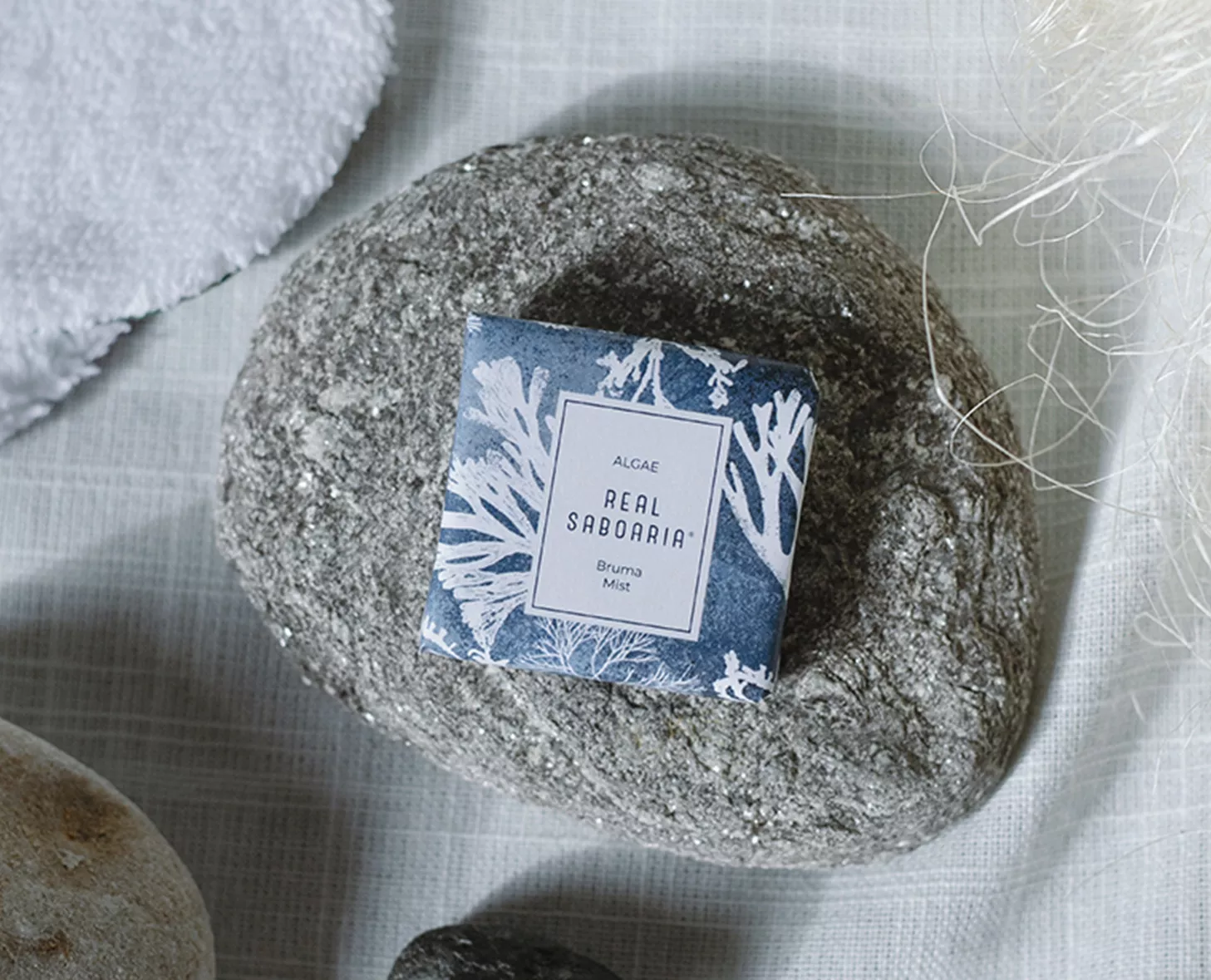 Una scatola di sapone blu e bianca con la scritta "Real Saborania" è appoggiata su una pietra grigia liscia, circondata da texture naturali tra cui tessuto morbido e spago.