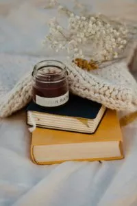 Eine gemütliche Dekoration mit einer gestrickten Decke, gestapelten Büchern und einer kleinen Kerze im Glas, mit zarten weißen Blumen im Hintergrund.