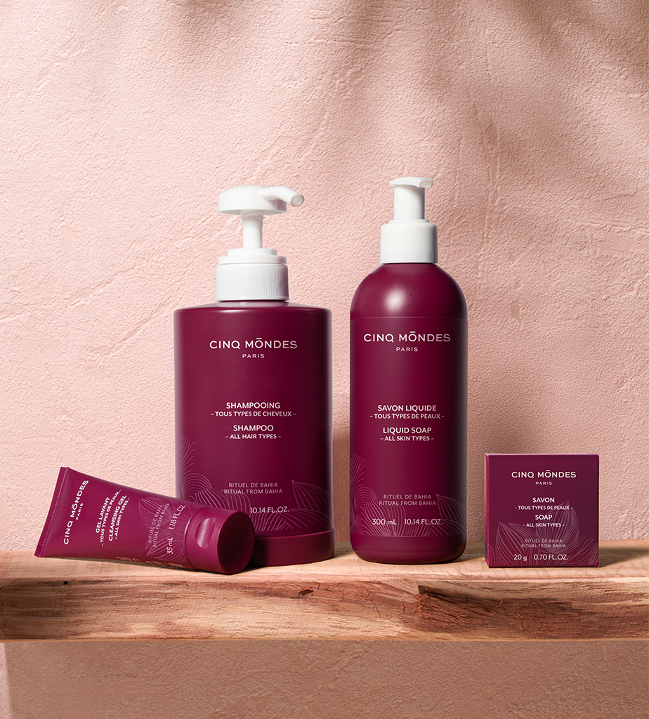 Uma coleção de produtos para cabelos e cuidados corporais da Cinq Mōndes Paris, incluindo um champô, sabonete líquido e uma pequena barra de sabonete, todos expostos contra um fundo rosa texturizado.