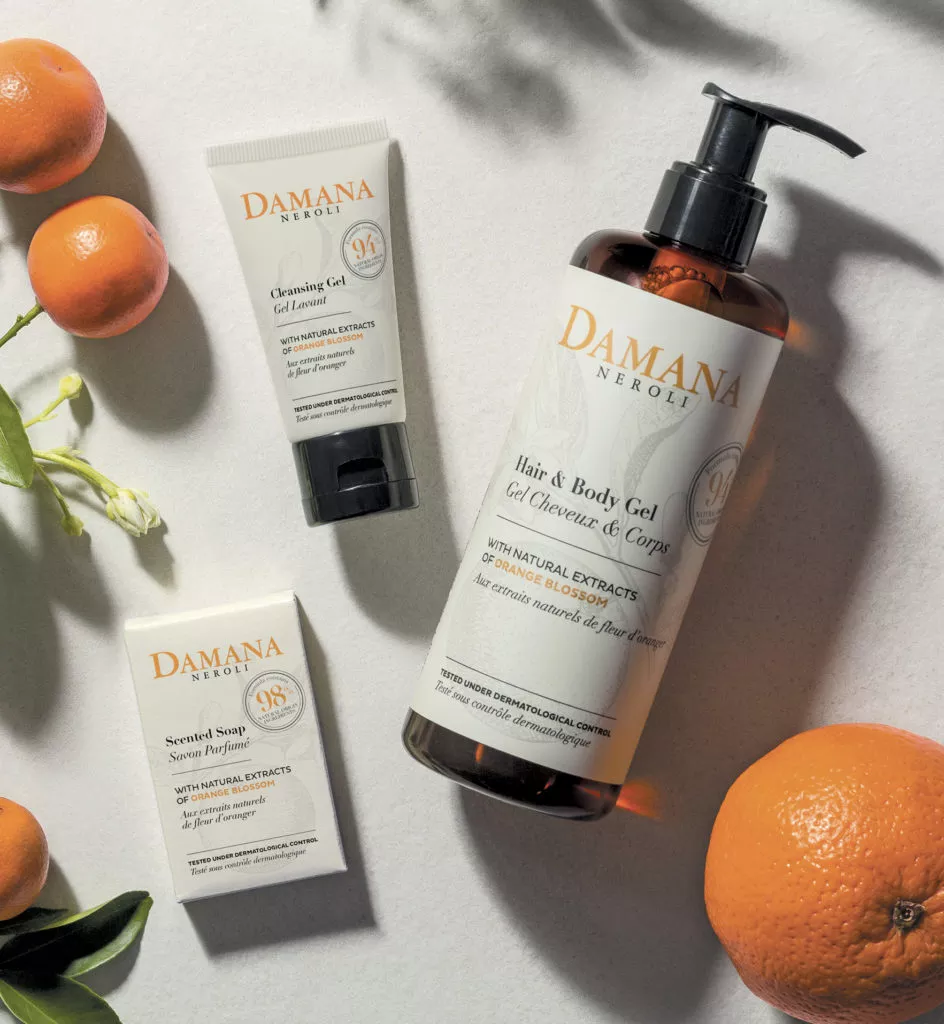 Eine Flasche Damana Neroli & Body Gel wird neben einer Tube Reinigungsgel und einem Stück duftender Seife präsentiert, im Hintergrund sind frische Orangen und grüne Blätter zu sehen. Die Produkte betonen natürliche Extrakte.