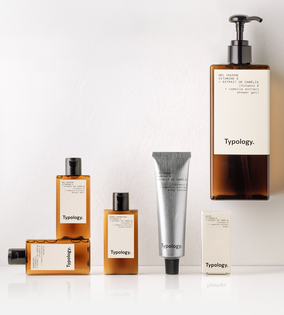 Eine Kollektion von Typology , angeordnet auf einer weißen Fläche, darunter ein Duschgel, Seren, eine Creme und ein Stück Seife, alle in eleganten braunen Flaschen und Behältern mit minimalistischen Etiketten.