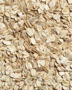 Uma imagem em grande plano de flocos de aveia, mostrando a sua forma oval achatada e cor esbranquiçada. Os flocos estão espalhados e densamente agrupados.