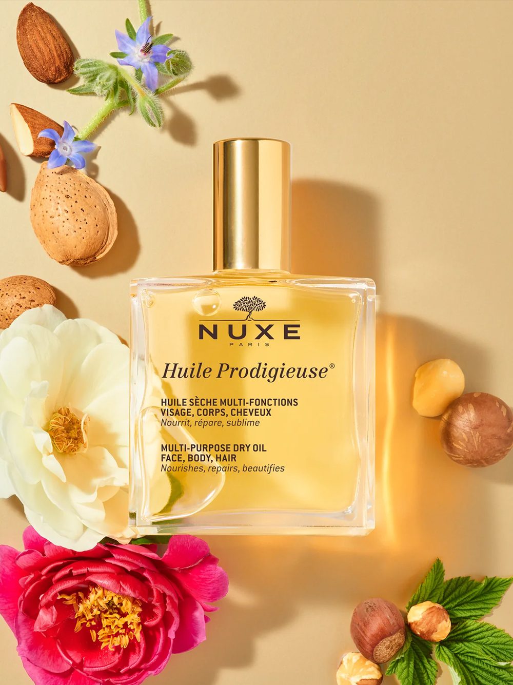 Eine Flasche NUXE Huile Prodigieuse Mehrzweck-Trockenöl, umgeben von bunten Blumen, Mandeln und Blütenblättern auf goldenem Hintergrund.