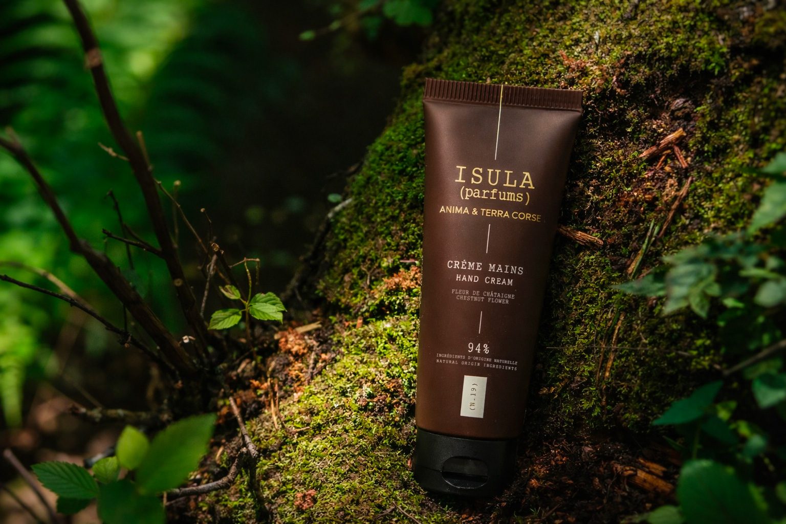 Un tubetto di crema Isula poggia su un tronco ricoperto di muschio circondato da fogliame verde. La confezione è marrone con scritte dorate, a indicare gli ingredienti naturali contenuti nel prodotto.