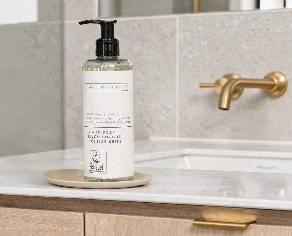 Eine Flasche Flüssigseife mit der AufschriftBaltic Bliss” steht auf einem kleinen beigen Teller auf einer Marmorarbeitsplatte, im Hintergrund ist ein stilvoller goldener Wasserhahn zu sehen. Die Flasche verfügt über einen Pumpverschluss und ist laut Aufschrift zu 100 % aus recycelten Materialien hergestellt.