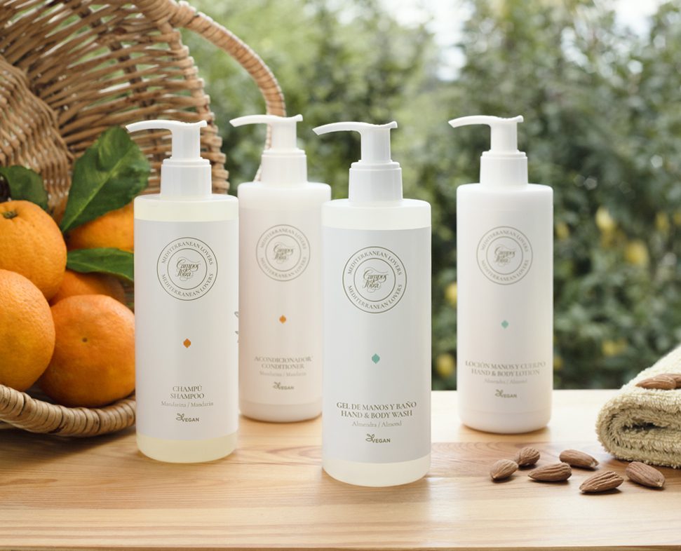 Una collezione di tre flaconi di prodotti per la cura dei capelli e del corpo disposti su una superficie di legno, con arance e mandorle sullo sfondo. I flaconi hanno un design minimalista bianco e presentano etichette di forma circolare.