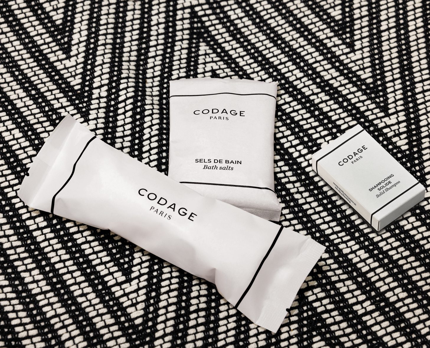 Drei Badezusätze CODAGE , darunter eine kleine Packung Badesalz und ein Stück Shampoo, präsentiert auf einem gemusterten schwarz-weißen Hintergrund.