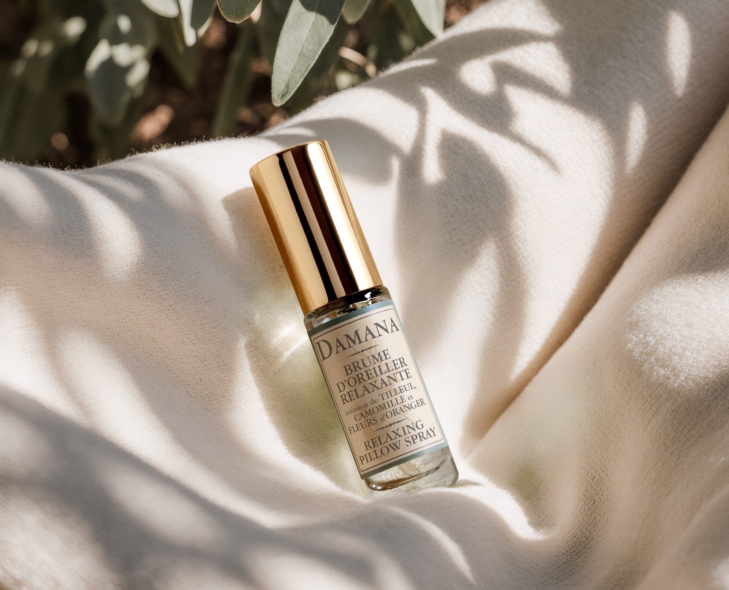Una piccola bottiglia di vetro di Damana Relaxing Pillow Spray con tappo dorato appoggiata su un tessuto chiaro, circondata da morbido fogliame verde.