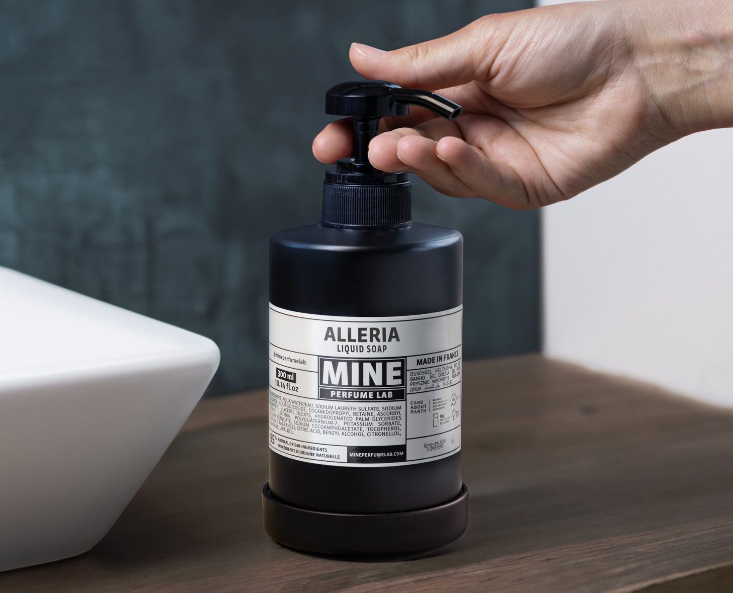 Uma mão segura um frasco com bomba de sabonete líquido Alleria com a etiqueta «MINE» sobre uma superfície de madeira, ao lado de um lavatório branco. O frasco apresenta um design moderno com uma etiqueta preta e branca.