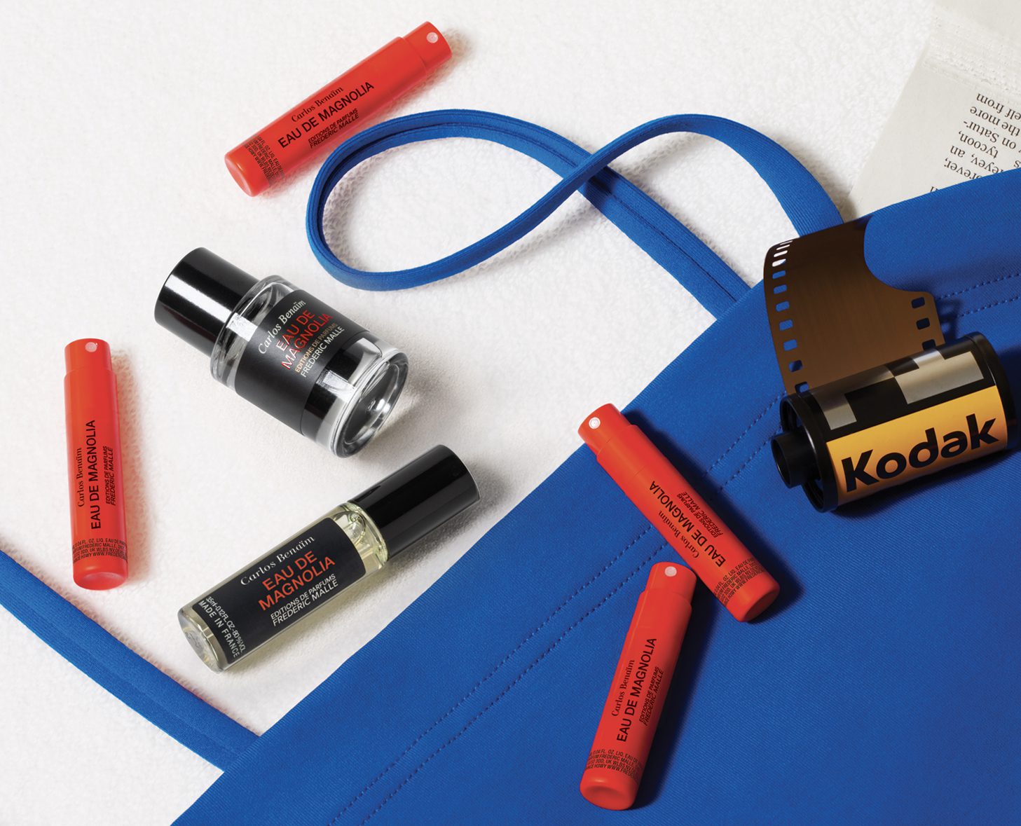 Una borsa blu circondata da vari cosmetici, tra cui rossetti rossi e un flacone di profumo, insieme a un rullino Kodak.