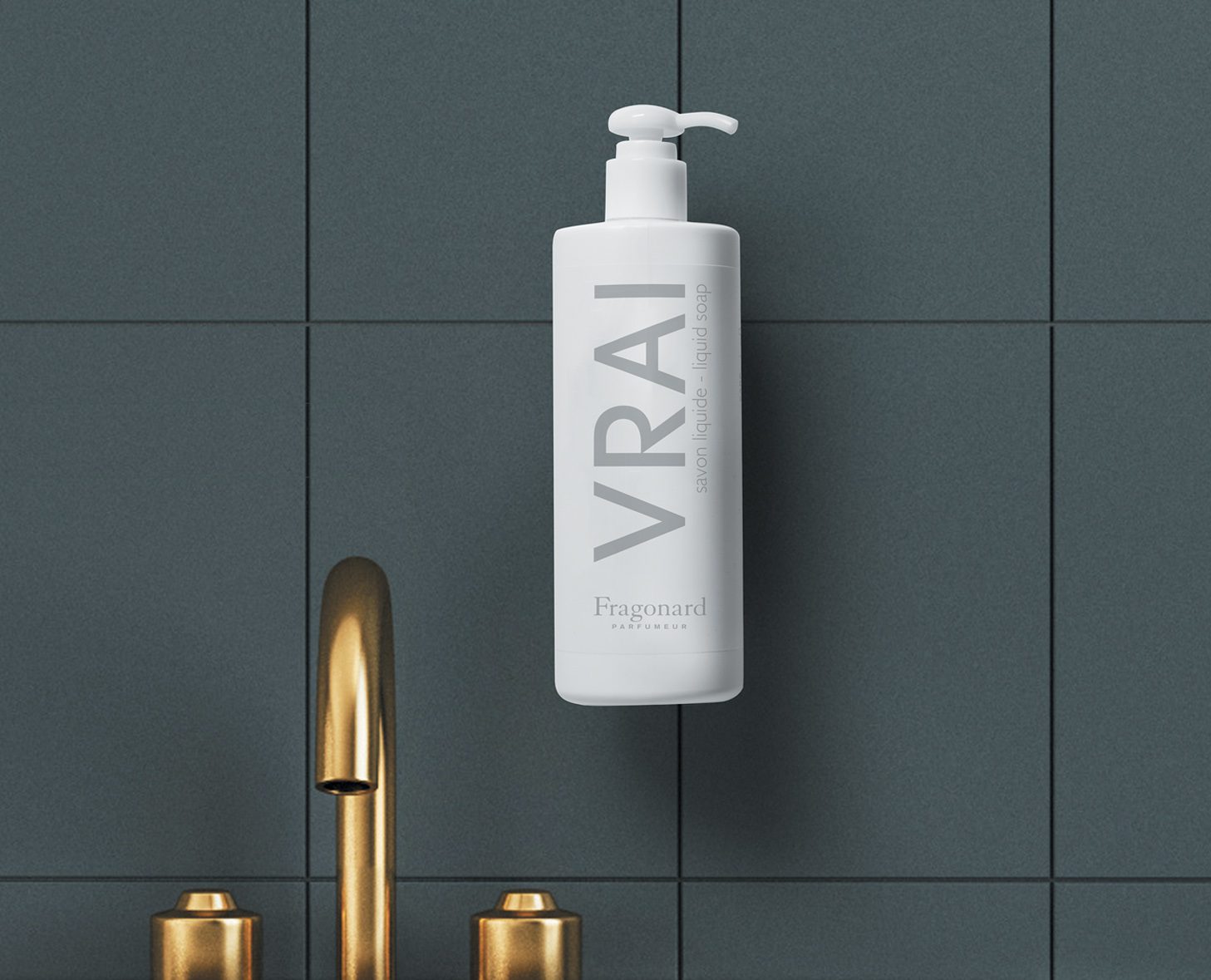 Un flacone bianco con pompa etichettato "VRAI" dal design minimalista, montato su una parete piastrellata scura accanto a un rubinetto dorato.