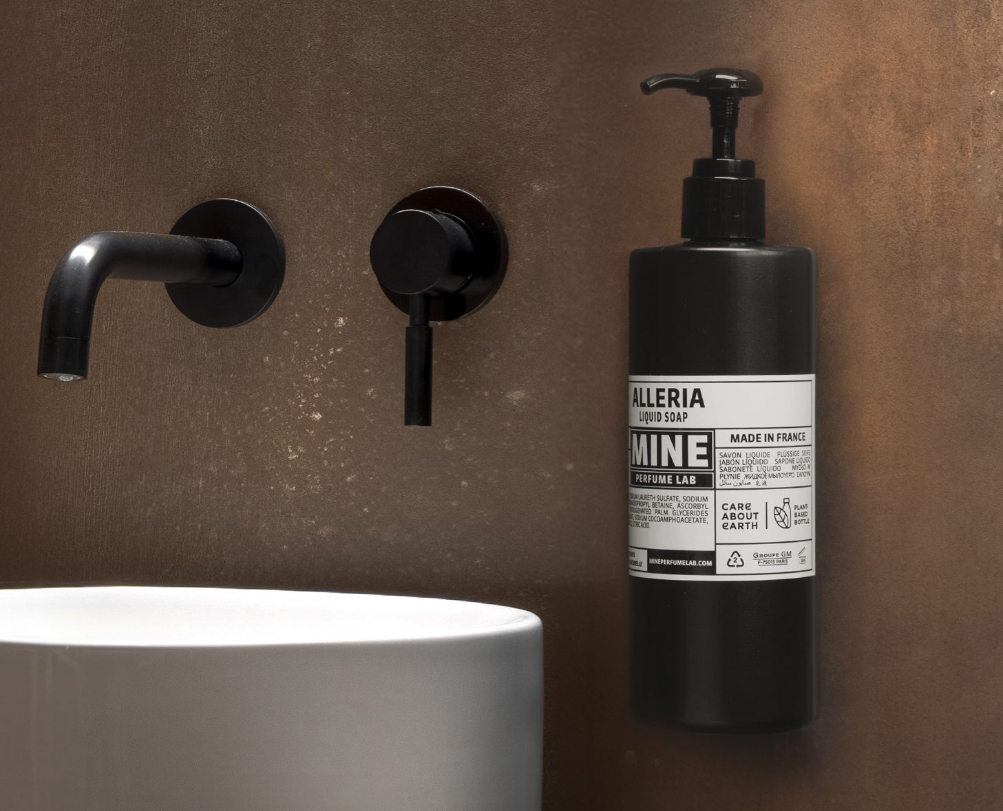 Um elegante ambiente de casa de banho com um dispensador de sabonete líquido preto com a marca «Alleria» montado numa parede texturizada, ao lado de torneiras pretas elegantes e um lavatório redondo branco.