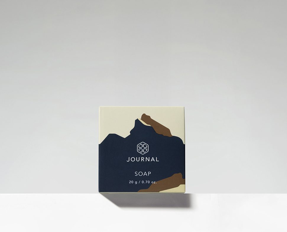 Una scatola di sapone minimalista con forme astratte marroni e crema su sfondo beige, con la parola "SOAP" e il nome del marchio "JOURNAL" ben visibili.