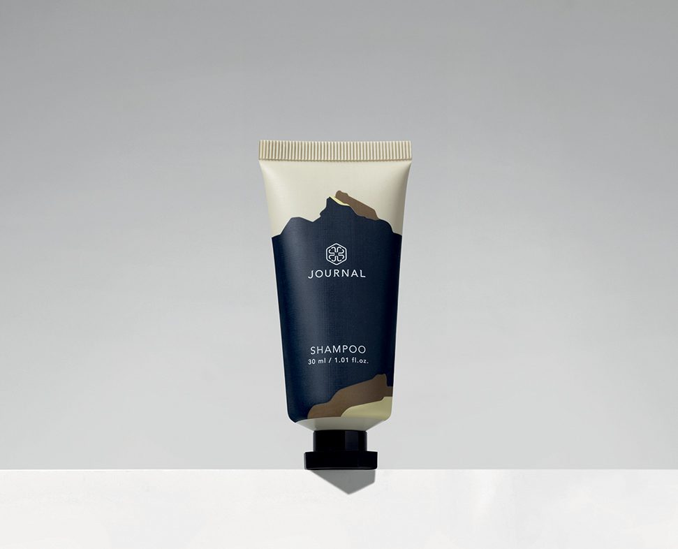 Un piccolo tubetto di shampoo con l'etichetta "JOURNAL" dal design minimalista, caratterizzato da una combinazione di colori blu scuro e crema su uno sfondo grigio semplice.