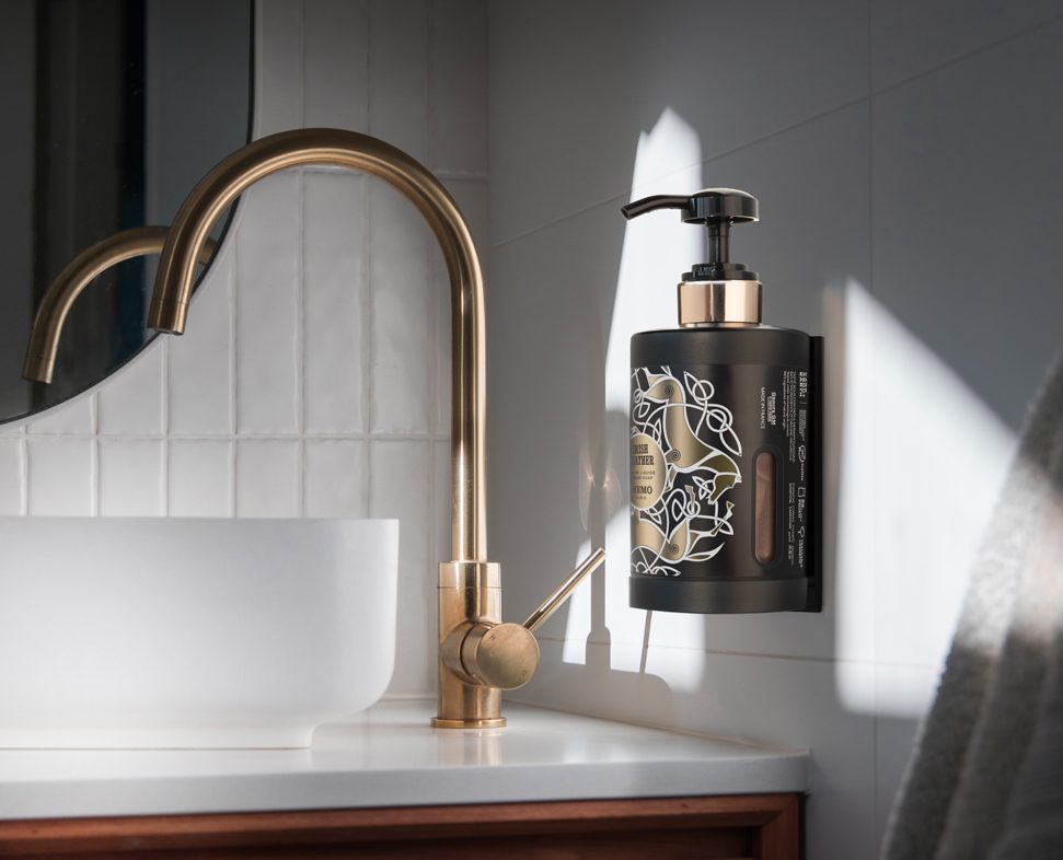 Ein schwarzer Flüssigseifenspender mit Pumpe ist an einer Badezimmerwand neben einem goldenen Wasserhahn und einem weißen Waschbecken angebracht und wird von natürlichem Licht beleuchtet.