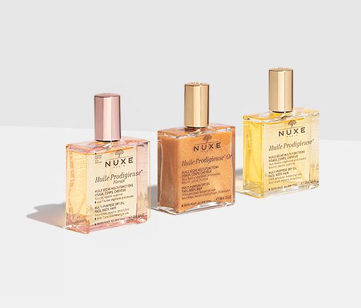 Três frascos de óleos NUXE Huile Prodigieuse, com líquido rosa, dourado e amarelo, expostos sobre um fundo claro. Cada frasco tem um rótulo e uma tampa dourada.