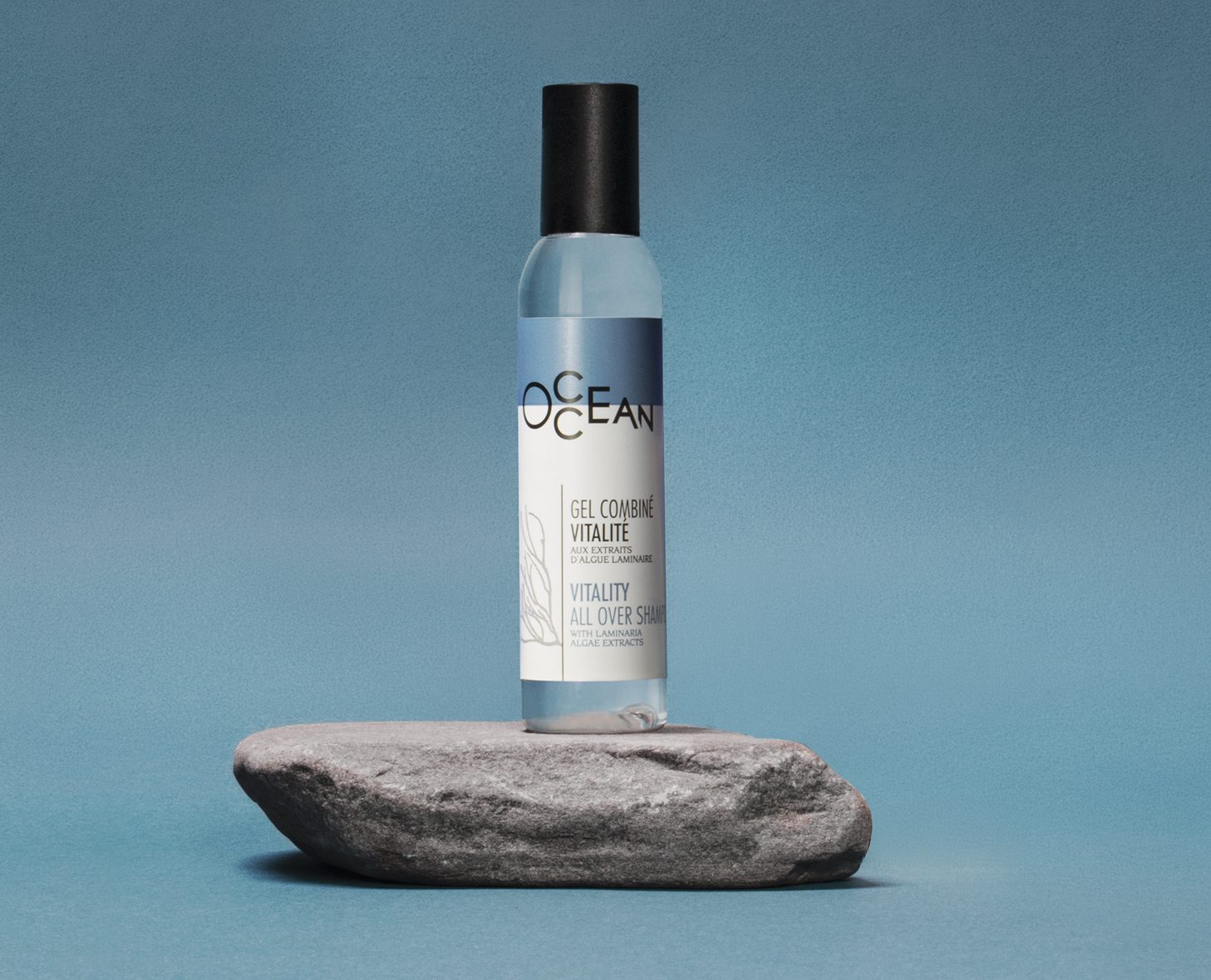 Una bottiglia di gel energizzante "OCEAN" è posizionata in verticale su una roccia grigia su sfondo blu. L'etichetta presenta un design minimalista con illustrazioni botaniche.