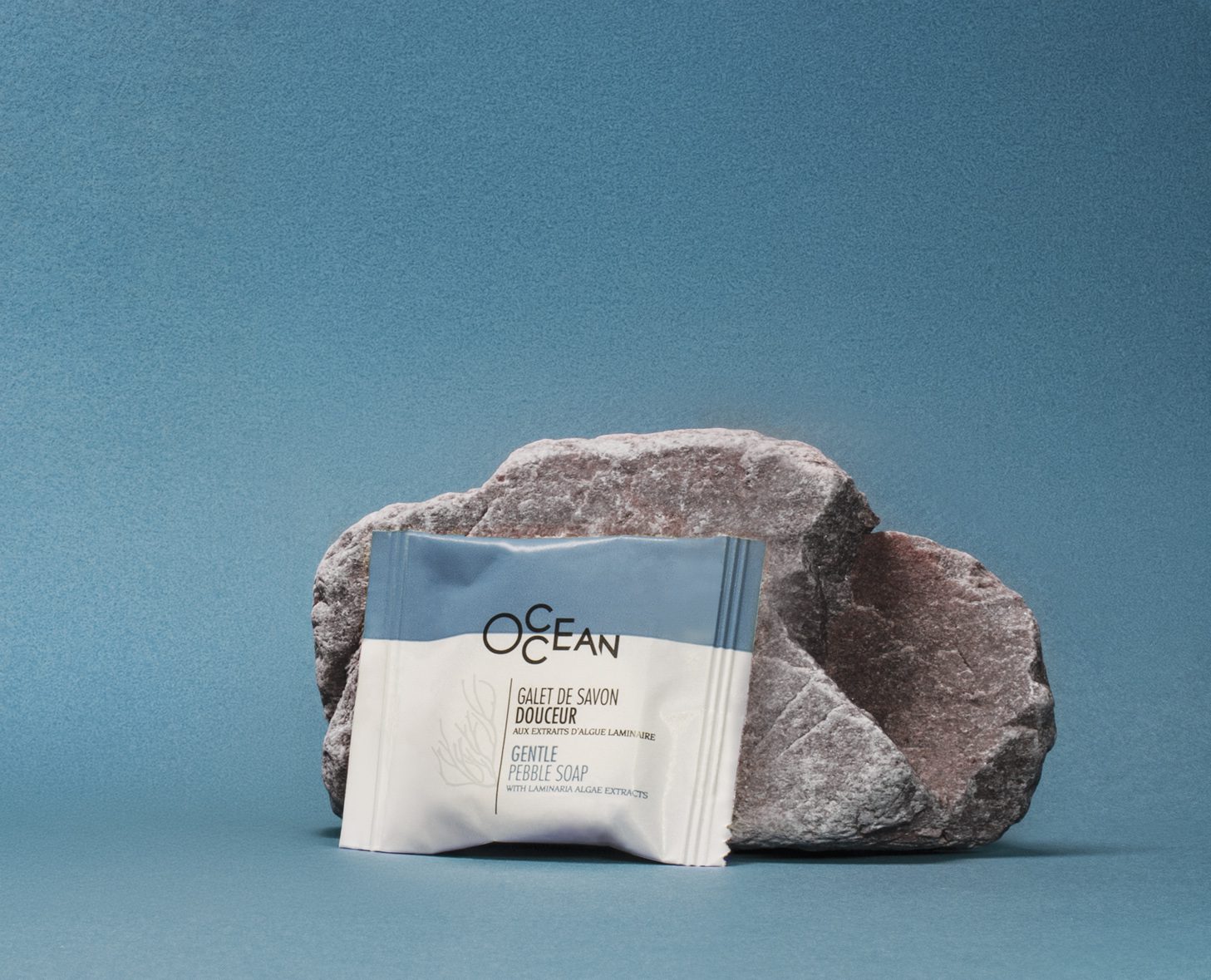 Una saponetta delicata a forma di ciottolo è posizionata accanto a una pietra ruvida su uno sfondo blu uniforme, mettendo in risalto la confezione con l'etichetta "OCEAN".