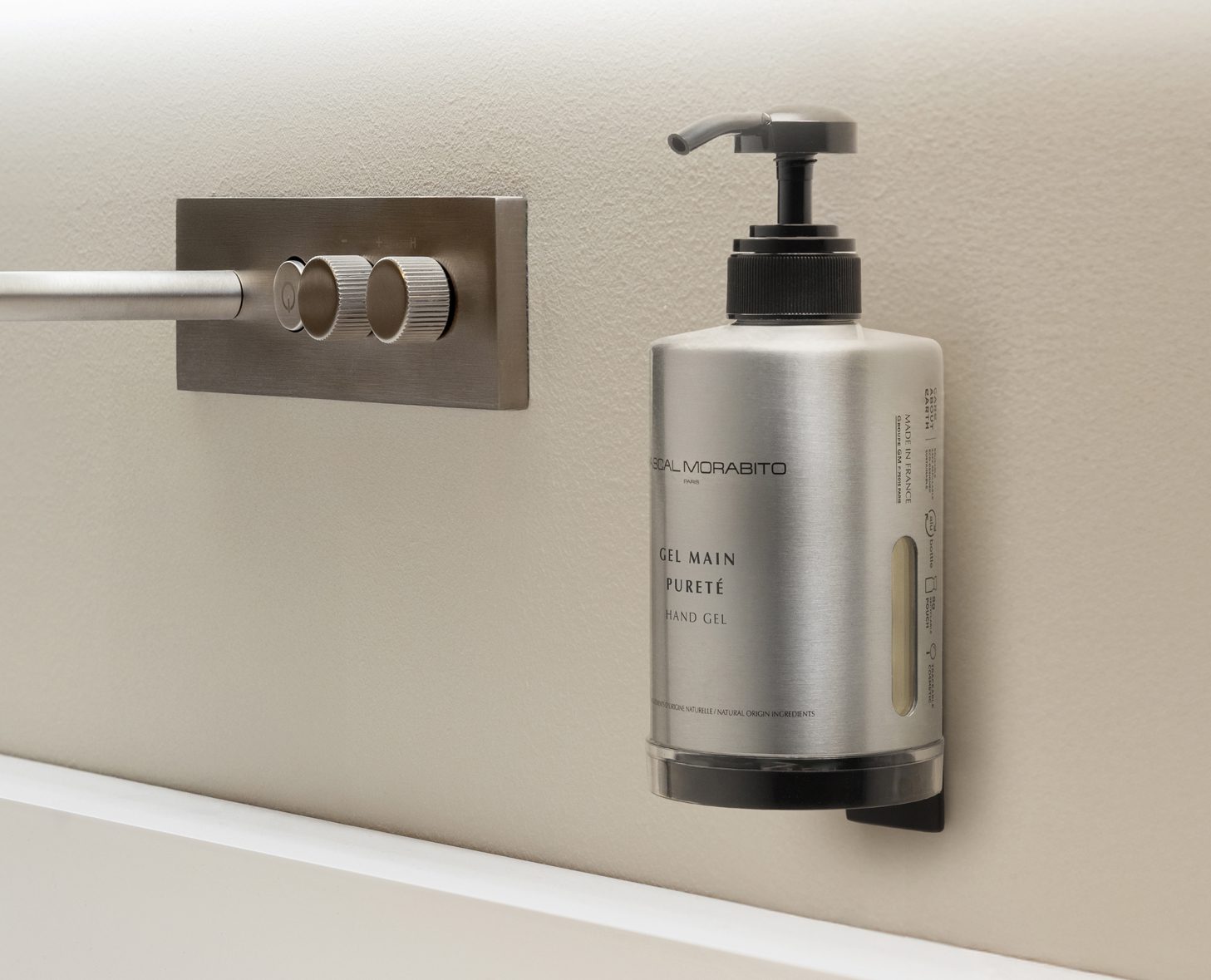 Ein Edelstahlspender für Handgel, der an einer Wand neben einem Regler für die Wassertemperatur angebracht ist. Der Spender verfügt über einen Pumpkopf und eine deutlich gekennzeichnete Inhaltsangabe.