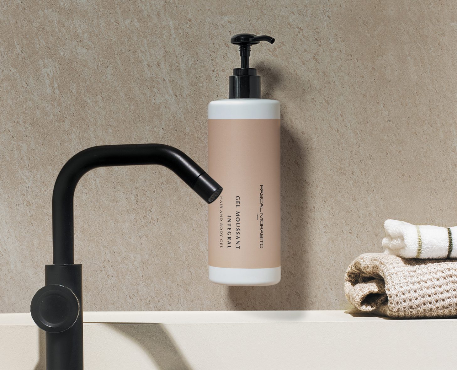 Eine Pumpflasche mit Reinigungsgel ist an einer hellen Steinwand neben einem schwarzen Wasserhahn und einem gefalteten cremefarbenen Handtuch angebracht.