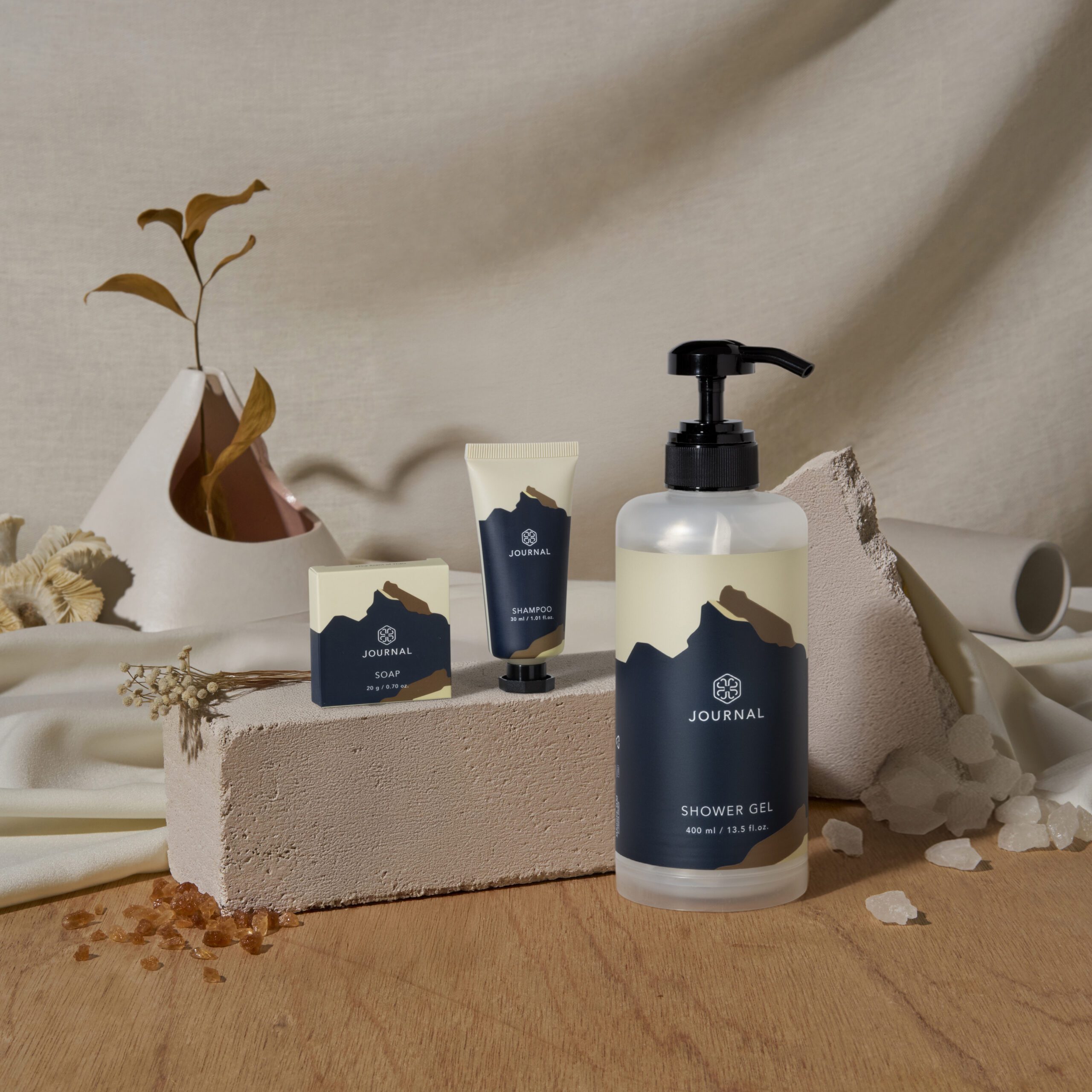 Una collezione di prodotti minimalisti per la bellezza e la cura personale disposti su una superficie strutturata, tra cui un flacone di bagnoschiuma, un tubetto di shampoo e una saponetta, su uno sfondo neutro caratterizzato da forme astratte e piante essiccate.