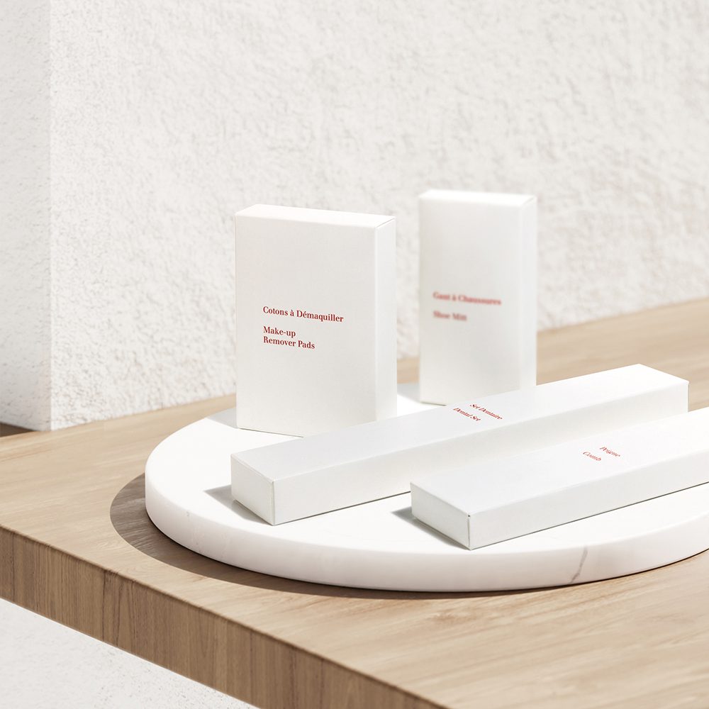 Una exposición minimalista de envases blancos para discos desmaquillantes y productos para el cuidado de la piel, con sencillas etiquetas de texto en rojo, dispuestos sobre una superficie redonda de mármol frente a una pared blanca con textura.