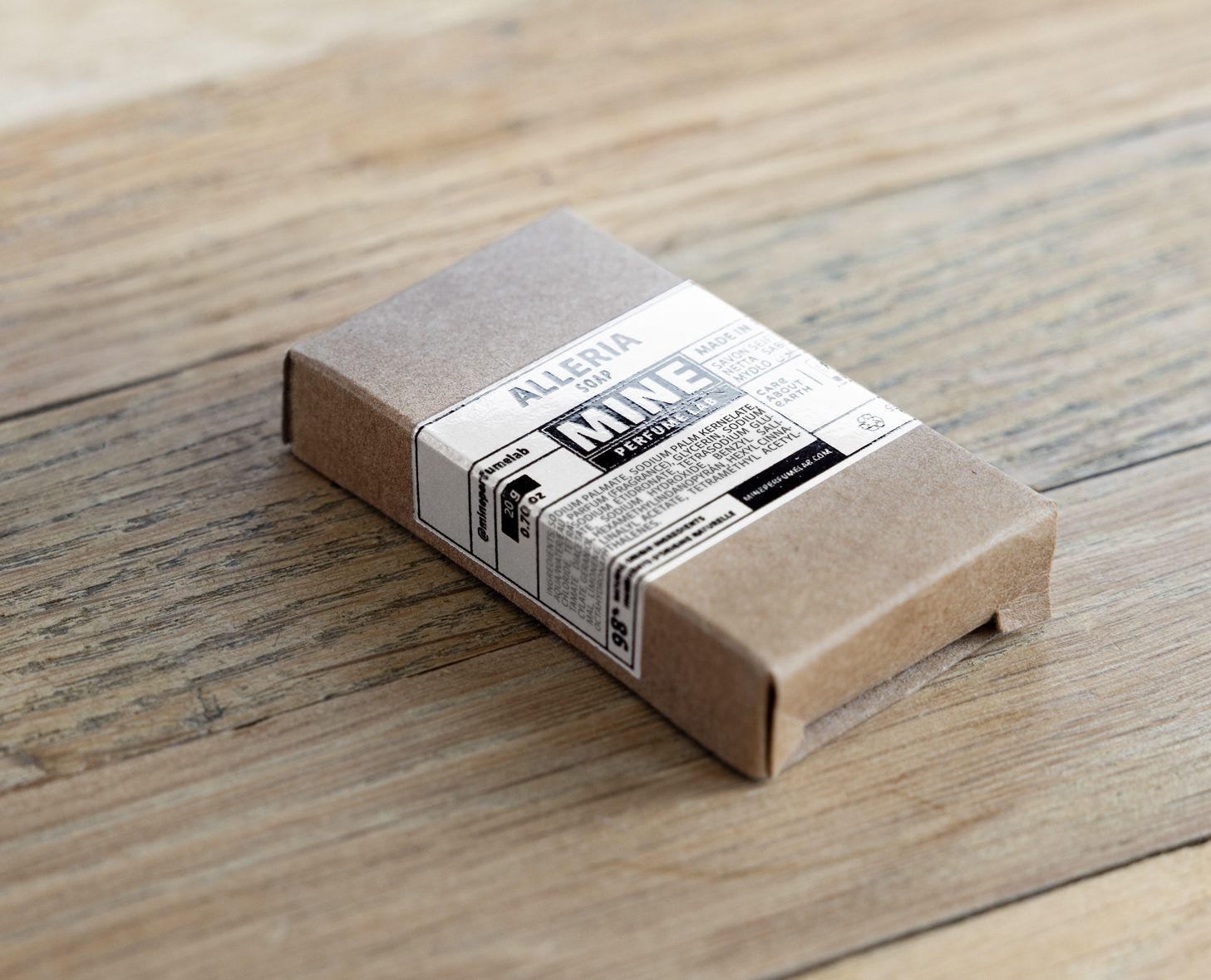 Uma caixa de cartão com uma etiqueta minimalista a preto e branco, apoiada numa superfície de madeira. A etiqueta apresenta um texto que inclui o nome da marca «Alleria» e menciona «MINE».