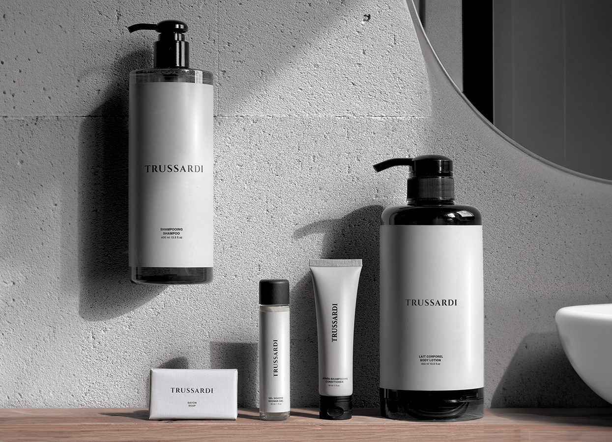 Eine minimalistische Anordnung von Trussardi auf einer Holzoberfläche vor einer Betonwand, mit Flaschen und Tuben in verschiedenen Größen.
