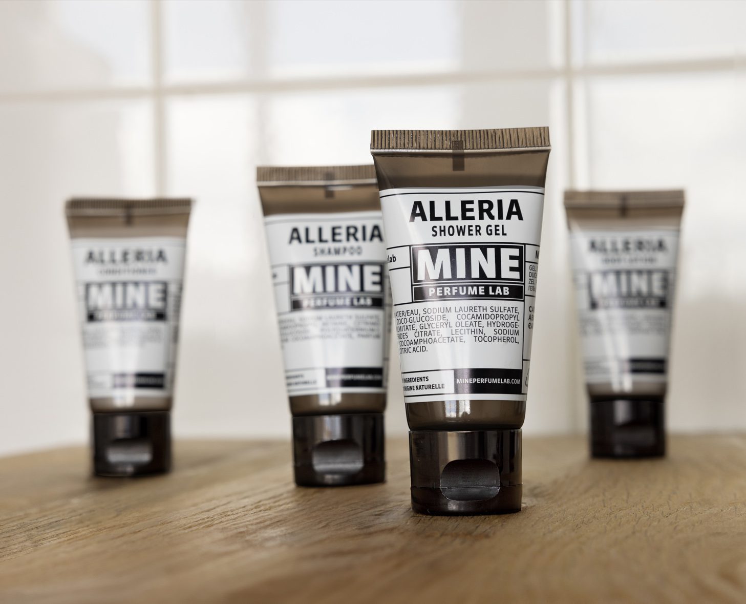 Quatro tubos de gel de banho e champô Alleria dispostos sobre uma superfície de madeira, com um design limpo e moderno e rótulos em preto e branco. O fundo apresenta azulejos de cor clara, criando um ambiente neutro.