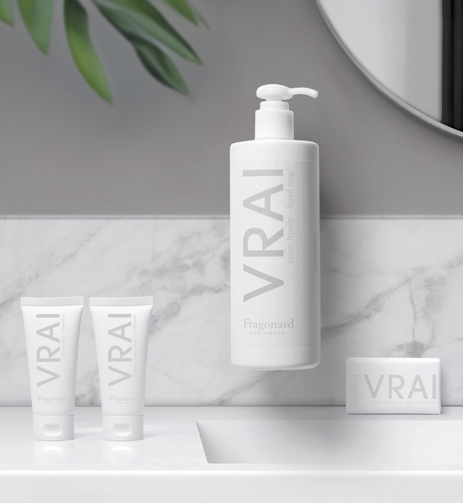 Una scena minimalista in bagno con vari prodotti per la cura della pelle con l'etichetta "VRAI", tra cui un grande flacone di sapone liquido, due tubetti e una saponetta, su uno sfondo di marmo e uno specchio circolare.