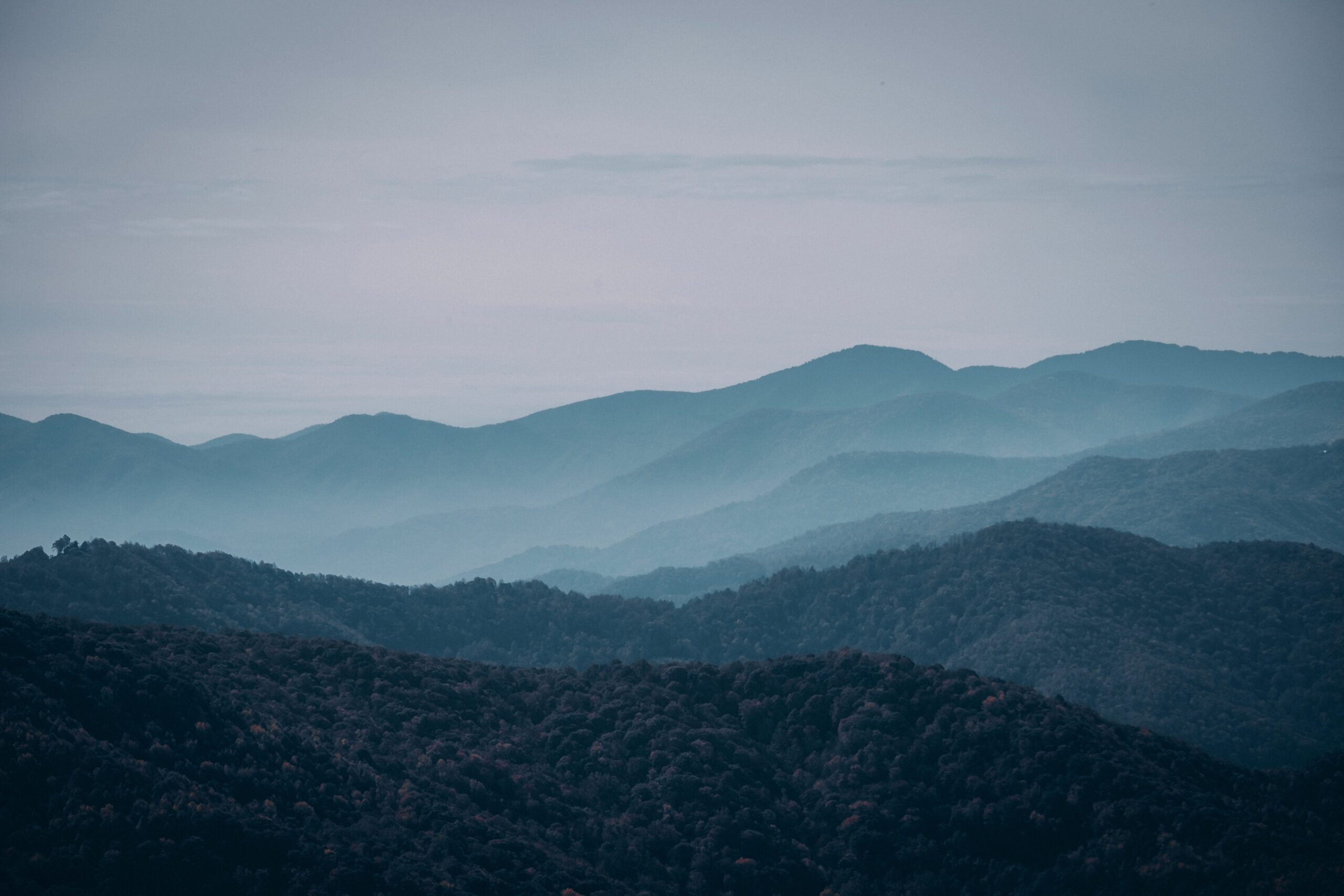 Una vista panoramica di montagne stratificate avvolte da una foschia nebbiosa, che creano un paesaggio sereno e suggestivo. Le diverse sfumature di blu e verde mettono in risalto i contorni naturali del terreno.
