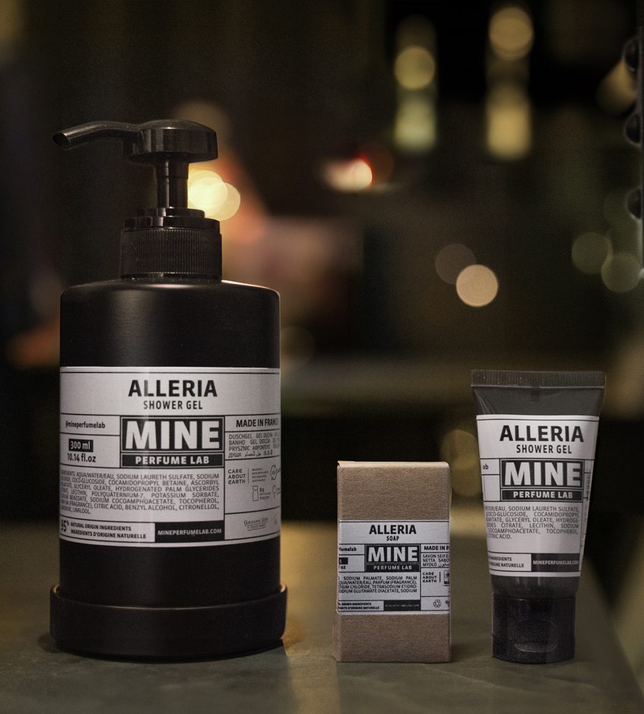 Um frasco preto de gel de banho Alleria ao lado de um pequeno tubo e uma caixa, todos com rótulos minimalistas com a palavra «MINE» em destaque. O fundo está suavemente desfocado com iluminação quente.