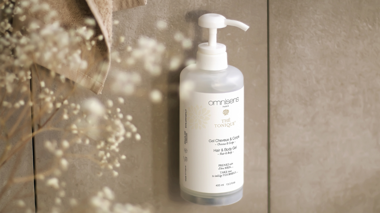 Omnisens Paris „Thé Tonique“ Haar- und Körpergel-Flasche an einer gefliesten Badezimmerwand mit zarten floralen Elementen im Vordergrund.