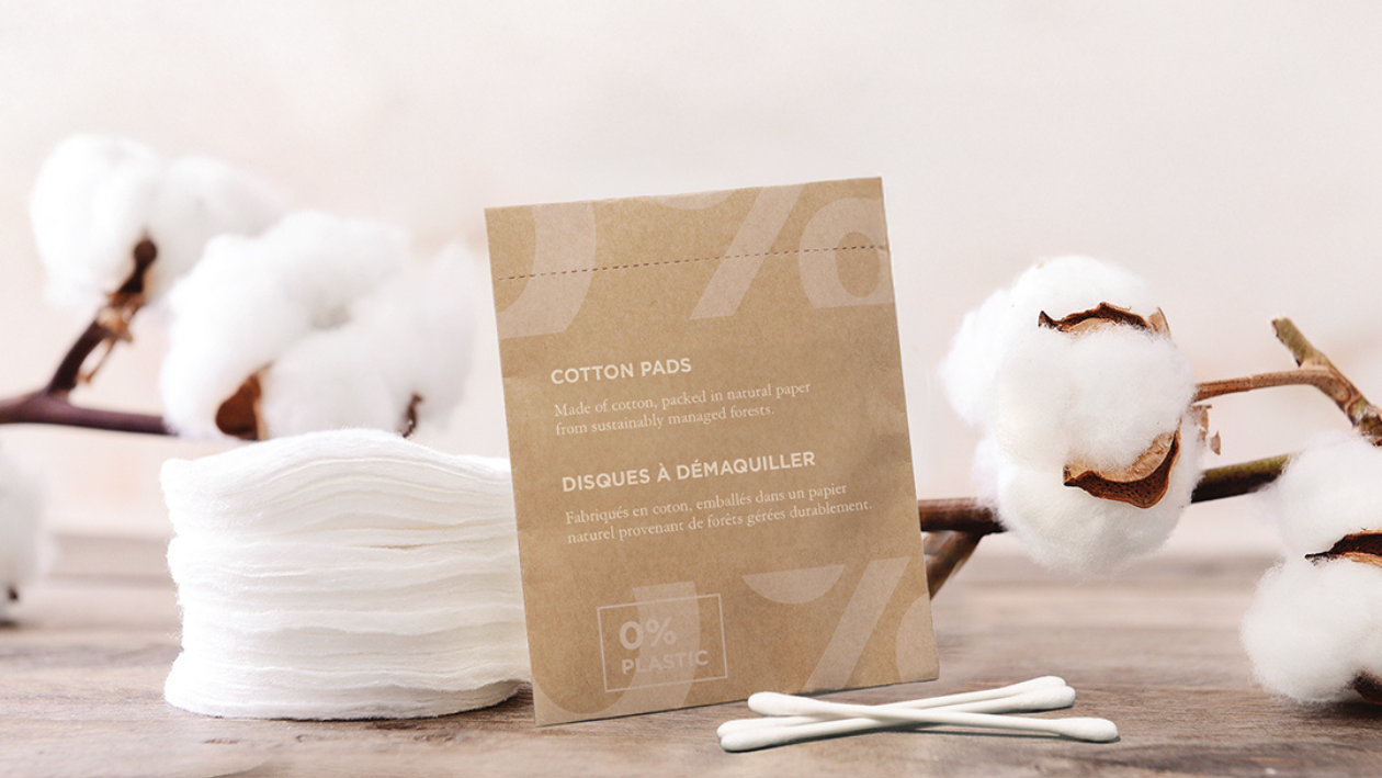 Umweltfreundliche Wattepads und Wattestäbchen in einer natürlichen Verpackung mit der Aufschrift „0 % Kunststoff“, präsentiert mit Baumwollblüten