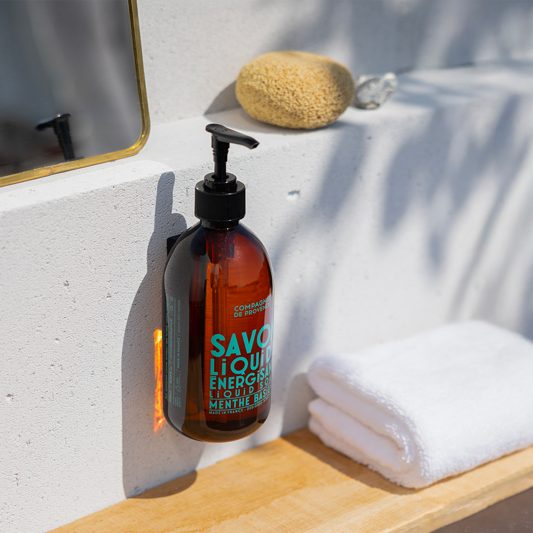 Compagnie de Provence Flüssigseifenflasche mit Pumpe, angebracht an einem Waschbecken, neben einem Handtuch und einem Schwamm in einem sonnendurchfluteten Badezimmer
